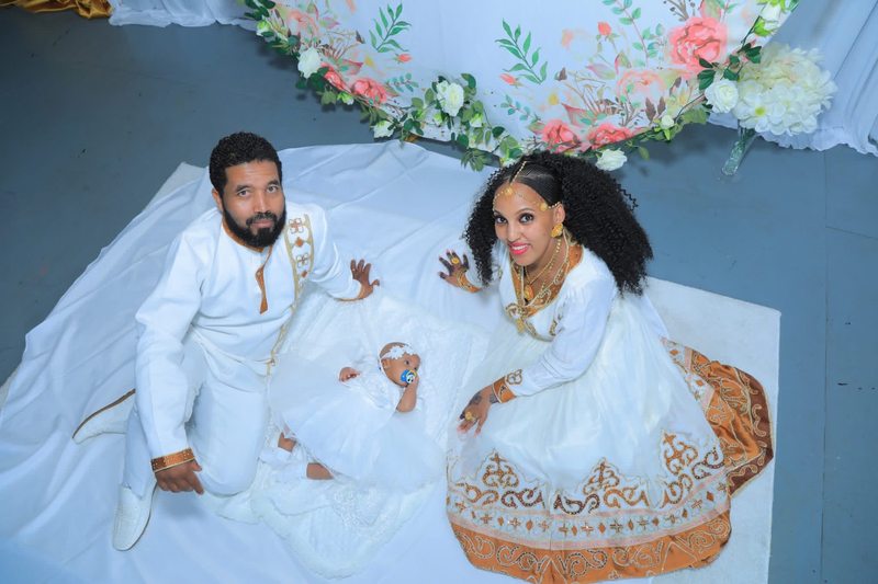 Parents seated beside their infant on a white blanket beneath floral decorations